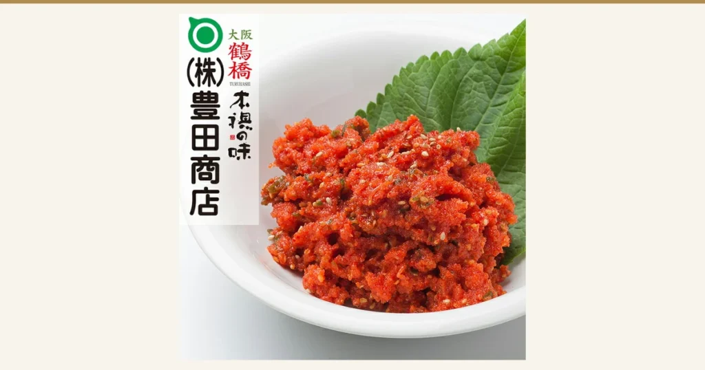 白い浅鉢に盛った、ごまの粒が見える赤いまかない明太子（230g）1パックと、後ろに添えた緑の葉の商品写真。左上に店舗が画像に入れた表示：「(株)豊田商店」のロゴ、「大阪 鶴橋 TURUHASHI」、「本場の味」