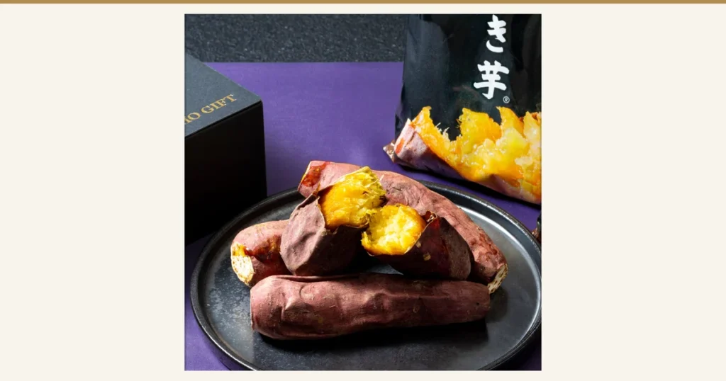 Product photo on a purple cloth: baked sweet potatoes with the cut flesh showing yellow on a black plate, with a black bagged product and a black box behind. Partial text visible: “き芋” on the bag and “GIFT” on the box (Zaiho frozen baked sweet potatoes, Beniharuka)