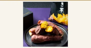 紫の布の上で、黒い皿に盛った焼き芋（割った断面は黄色）。奥に黒い袋入りの商品と黒い箱が写る商品写真。袋の一部に「き芋」、箱に「GIFT」の文字が見える（財宝 冷凍焼き芋 紅はるか）