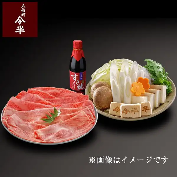 黒い背景に、丸皿に広げた黒毛和牛すき焼き用の肩・肩ロース薄切り肉と、白菜・長ねぎ・春菊・生しいたけ・花人参・焼き豆腐・丁字麩などを盛った器、すき焼割下の瓶が並んだ商品画像。店舗ロゴ「人形町今半」と「※画像はイメージです」の文字入り