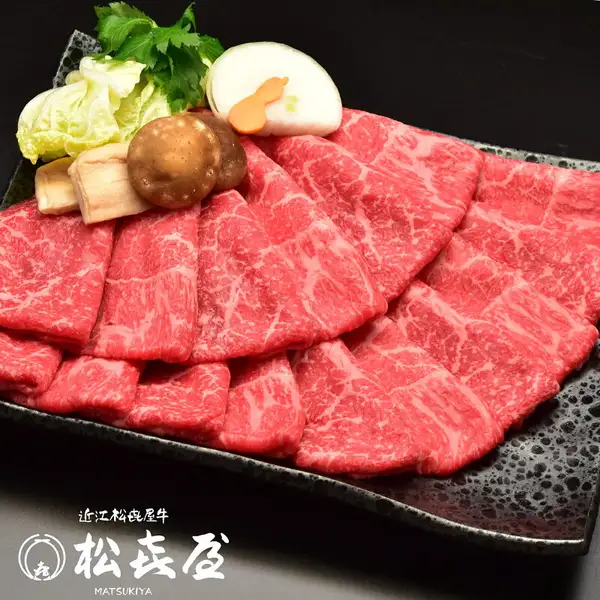 【特上】近江牛 すき焼き用 モモ・バラ1kgのスライス肉を黒い角皿に扇状に盛り付けた写真。白菜・三つ葉・しいたけ・長ねぎ・にんじんなどを添えているが、これらの野菜・きのこは商品に含まれない。画像内に店舗ロゴ「近江松喜屋牛」「松喜屋 MATSUKIYA」の文字あり。