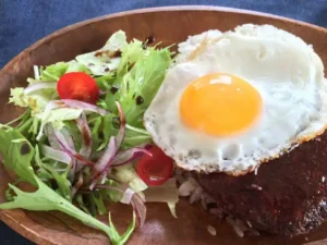Loco moco-style serving example of ゆうぜん 100% beef, additive-free hamburg steak on a wooden plate: the hamburg steak glazed with dark brown sauce on rice, topped with a fried egg, with a salad of lettuce, mizuna, red onion and 2 cherry tomatoes (the rice, egg and salad are not included with the product). There is no text in the image