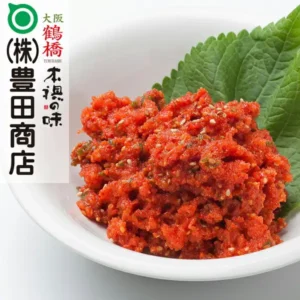 白い浅鉢に盛った、ごまの粒が見える赤いまかない明太子（230g）1パックと、後ろに添えた緑の葉の商品写真。左上に店舗が画像に入れた表示：「(株)豊田商店」のロゴ、「大阪 鶴橋 TURUHASHI」、「本場の味」