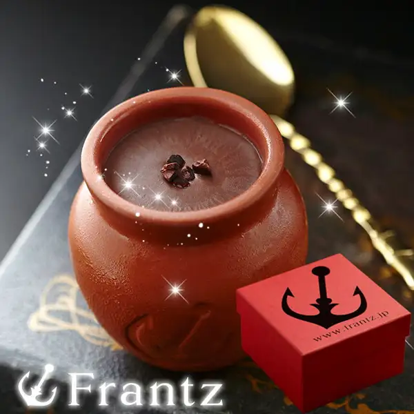 Photo of one chocolate pudding in a reddish-brown pot-shaped container (Kobe Frantz Kobe Mahou no Tsubo Pudding®, Chocolat, 4 pieces), with cacao nibs on top, a gold spoon and a small red box beside it. The shop logo "Frantz" is at lower left and the text "www.frantz.jp" is on the small box