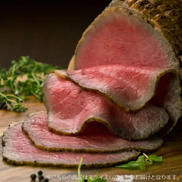 木板上摆放着表面烤出焦色的整块肉のいとう 仙台牛ローストビーフ（烤牛肉），以及从中切下的4片断面呈粉色的薄片（2片折叠重叠、2片平铺叠放），旁边摆有香草、绿叶菜和黑胡椒粒的摆盘示例照片（香草、绿叶菜和黑胡椒粒不包含在商品中）。图片下方有店铺放入图片中的文字「※こちらの商品はスライスし冷凍でのお届けとなります。」