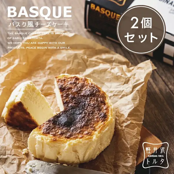 軽井沢トルタ バスク風チーズケーキの商品画像。表面が焦げ茶色に焼き色づいたホールのチーズケーキから扇形に1切れが切り出され、クリーム色の断面を見せて手前に置かれている。皺のあるクラフト紙の上、木のカッティングボードに置かれ、手前にナイフが写っている。画像には「BASQUE」「バスク風チーズケーキ」の文字、英文メッセージ「THE BASQUE CHEESE CAKE IS PRODUCT OF KARUIZAWA TORTA. WE HOPE YOU ARE HAPPY WITH OUR PRODUCTS. PEACE BEGIN WITH A SMILE.」、「2個セット」と書かれた丸いバッジ、「軽井沢トルタ KARUIZAWA TORTA」のロゴが焼き込まれている。