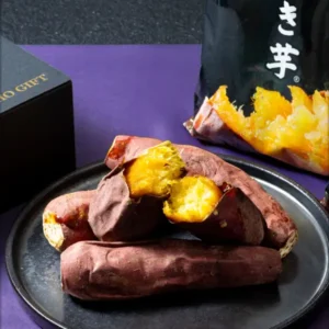 紫の布の上で、黒い皿に盛った焼き芋（割った断面は黄色）。奥に黒い袋入りの商品と黒い箱が写る商品写真。袋の一部に「き芋」、箱に「GIFT」の文字が見える（財宝 冷凍焼き芋 紅はるか）