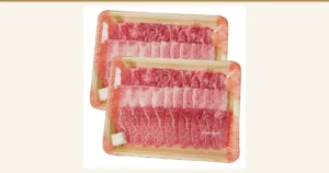 飛騨牛カルビ 焼肉用 メガ盛り1kg的商品照片：两个用透明薄膜包裹的木纹托盘中，摆放着带有红肉和白色脂肪的烤肉用薄切牛肉，托盘的一角附有小白块。照片右下角有店铺名的水印。