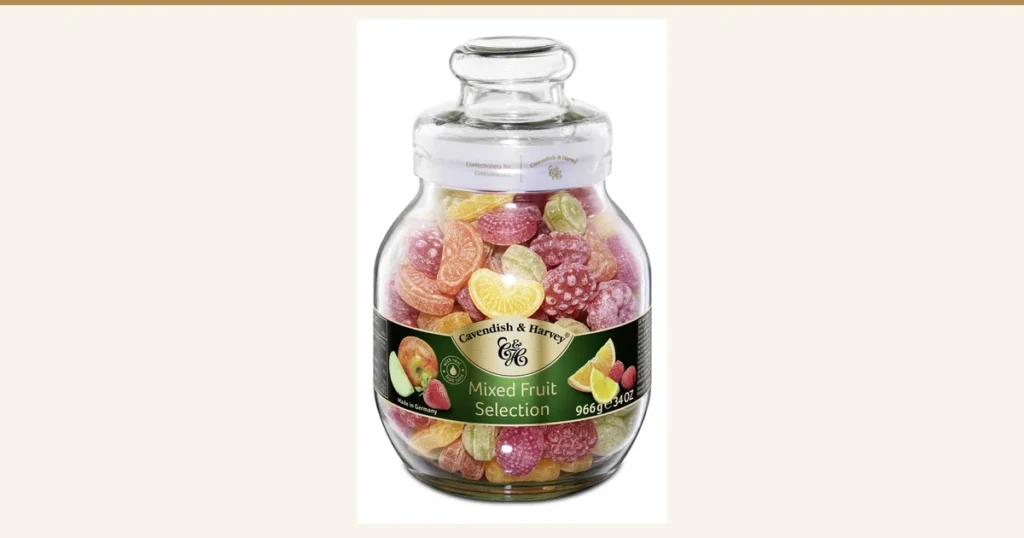 Product photo of the Cavendish & Harvey Mixed Fruit Candy Jar 966 g in a glass jar. A round jar with a clear lid is filled with colorful candies shaped like oranges, raspberries, lemons and other fruit. The green label on the jar shows the packaging print "Cavendish & Harvey", "Mixed Fruit Selection", "966g" and "Made in Germany".