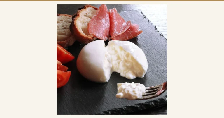 Product photo of a white round burrata (fresh mozzarella) cheese split open on a black slate plate, with a forkful of the filling (花畑牧場 Burrata Fresh Mozzarella Cheese, 70g×12 Pieces). Sliced cured ham, baguette slices and tomato wedges are also on the plate as a serving example and are not included with the product