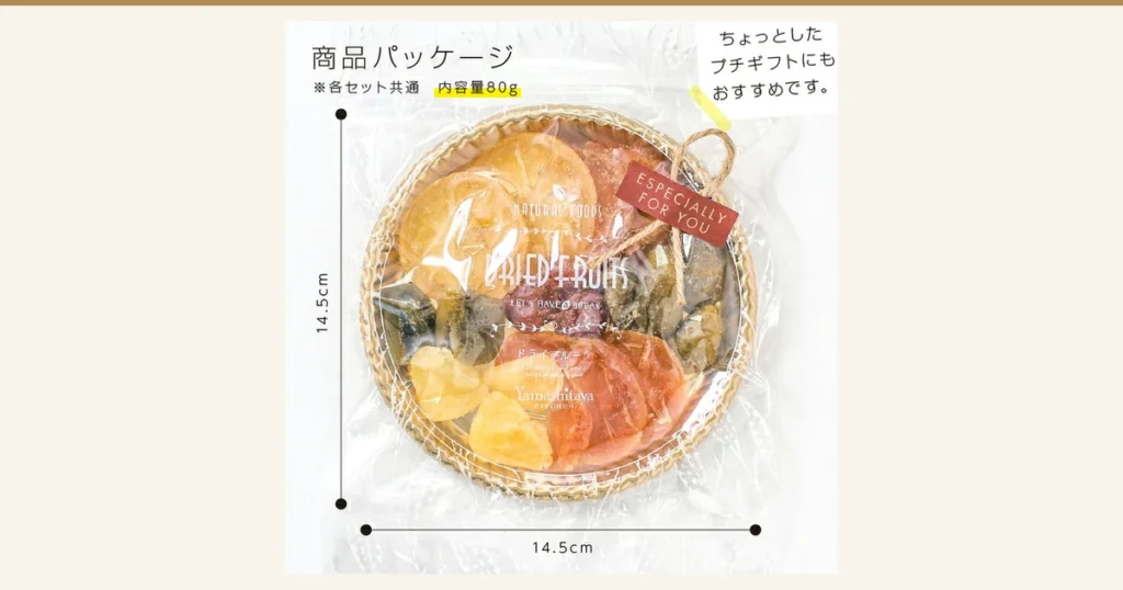 Package photo of Japan-grown Dried Fruit Tasting Assortment, 7 Kinds (Standard Set), 山下屋荘介: a round woven-basket-style tray inside a clear bag, with sliced dried fruit and yellow cubes arranged in a circle, tied with twine and a red tag, with dimension lines at the left and bottom of the bag. Text the shop placed in the image reads “商品パッケージ” (Product package), “※各セット共通 内容量80g” (Common to all sets, contents 80g) and “ちょっとしたプチギフトにもおすすめです。” (Also recommended as a small gift), with dimensions “14.5cm” and “14.5cm”. Printed on the bag: “NATURAL FOODS”, “DRIED FRUITS”, “ドライフルーツ”, “Yamashitaya KITCHEN”; on the tag: “ESPECIALLY FOR YOU”.