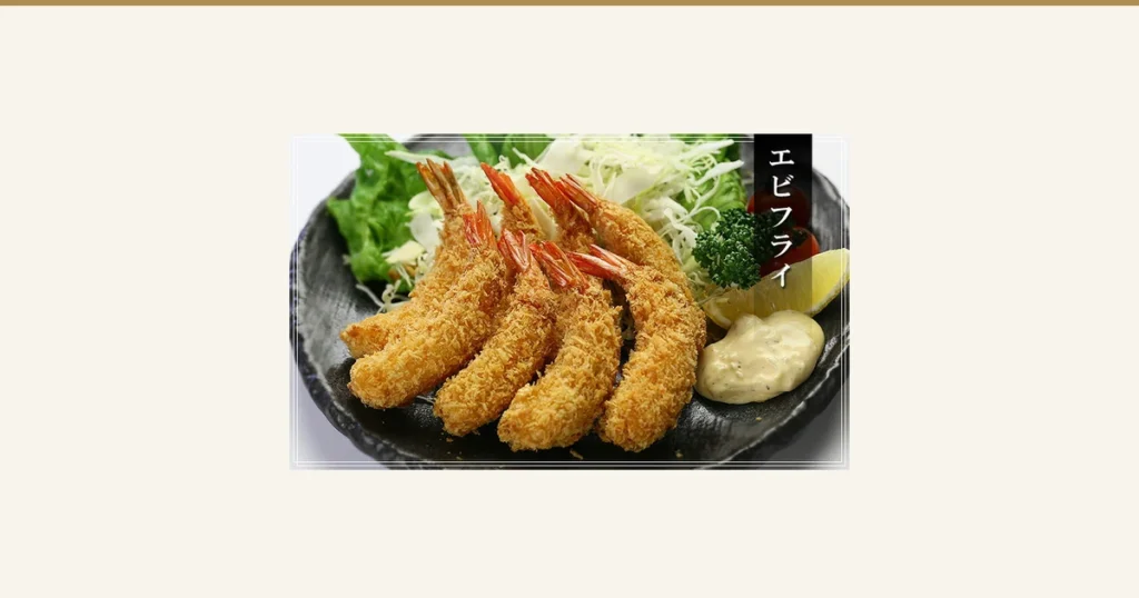 盛在黑色盘子里的炸虾摆盘示例照片，配有切丝卷心菜、生菜、欧芹、小番茄、柠檬和塔塔酱（甲羅組 天然えびフライ 8尾入）。店铺在图片中加入的标示「エビフライ」（炸虾）。配菜不包含在商品中