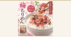 Featured image for Ume Chirimen Soft Furikake with Crunchy Ume (うめ海鮮). A serving-example product photo: a generous heap of furikake mixing chirimen (small dried sardines), red crunchy ume, hijiki, wakame and sesame sits on white rice in a whitish bowl, with some lifted by chopsticks. At the lower left is one clear bag of the product with a pink label; in the blurred background are a dish of a red food, green leaves and cherry-blossom decorations (the rice and the food in the background dish are not included with the product). The store’s own text in the image reads “梅ちりめん” (ume chirimen), “カリカリ梅使用” (made with crunchy ume) and “海鮮ふりかけ” (seafood furikake), and the bag label reads “自然の恵み 海の幸” (nature’s blessings, treasures of the sea), “梅” and “ちりめん”.