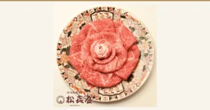 Product photo of Omi beef sukiyaki slices (loin, momo and bara) arranged in the shape of a rose on a round plate with a red, orange and black floral pattern. The store logo with the text “近江松喜屋牛” and “松喜屋 MATSUKIYA” appears in the image (shown by the store). Tokusen Omi Gyu Sukiyaki Beef 1kg, Matsukiya