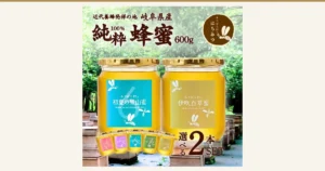 春日養蜂場（みつばち村）国产蜂蜜600g×2瓶套装的商品照片。以树林和木箱为背景，两瓶金色瓶盖的蜂蜜（标签为“初夏の里山蜜”“伊吹百草蜜”）并排而立，前方另有五瓶较小的瓶子。图片内为店铺自行加入的文字：“近代養蜂発祥の地 岐阜県産”“純粋100% 蜂蜜 600g”“選べる2本SET”及标志“みつばち村のはちみつ”。
