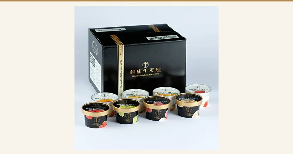 Product photo of a black box with 4 black cups with gold lids (ice cream) in the front row and 4 white cups with clear lids (sorbet) in the back row. The front lids carry the text “銀座プレミアムアイス”, the English names “TOCHIOTOME”, “MUSCAT”, “PEACH” and “CUSTARD VANILLA”, and pictures of fruit and other ingredients; the back lids show mango, pineapple, yuzu with honey, and strawberry. The box and cups carry the shop's logo “銀座千疋屋” and “Ginza Sembikiya since 1894” (a mark the shop placed in the image). Ginza Premium Ice & Sorbet, 8 Pieces (パティスリー銀座千疋屋)