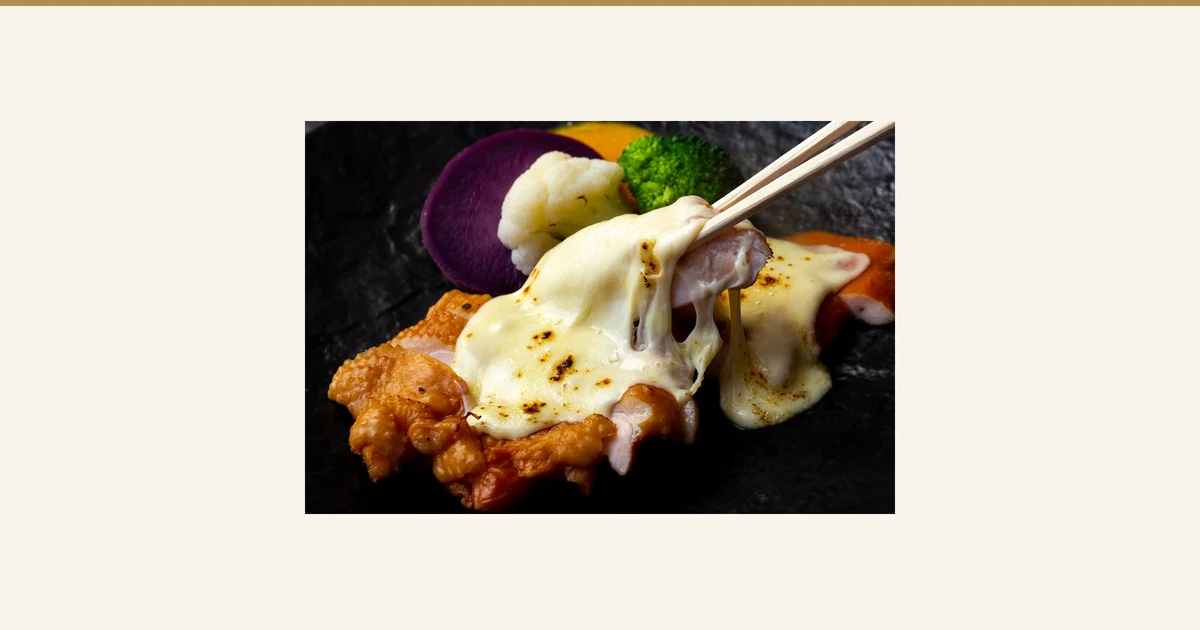 黒い石板風の皿にのせた燻製チキンにとろけたチーズを重ね、箸で持ち上げた盛り付け例の写真(純系名古屋コーチン 燻製4種セット)。皿の奥に紫いも・カリフラワー・ブロッコリー・かぼちゃが見える。チーズと野菜は商品に含まれません。