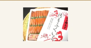 Product photo of 築地丸中 特上博多ふくいち粒々辛子明太子1kg (Tsukijimarunaka Tokujo Hakata Fukuichi Tsubutsubu Karashi Mentaiko 1kg): orange-tinted pink spicy mentaiko roe sacs in a white tray, laid out in three rows separated by green dividers, with the package lid laid over the right side of the tray. The lid is printed with “辛子明太子”, “博多ふくいち” and “素材と味へのこだわり。真心込めて作りました。” as package lettering.