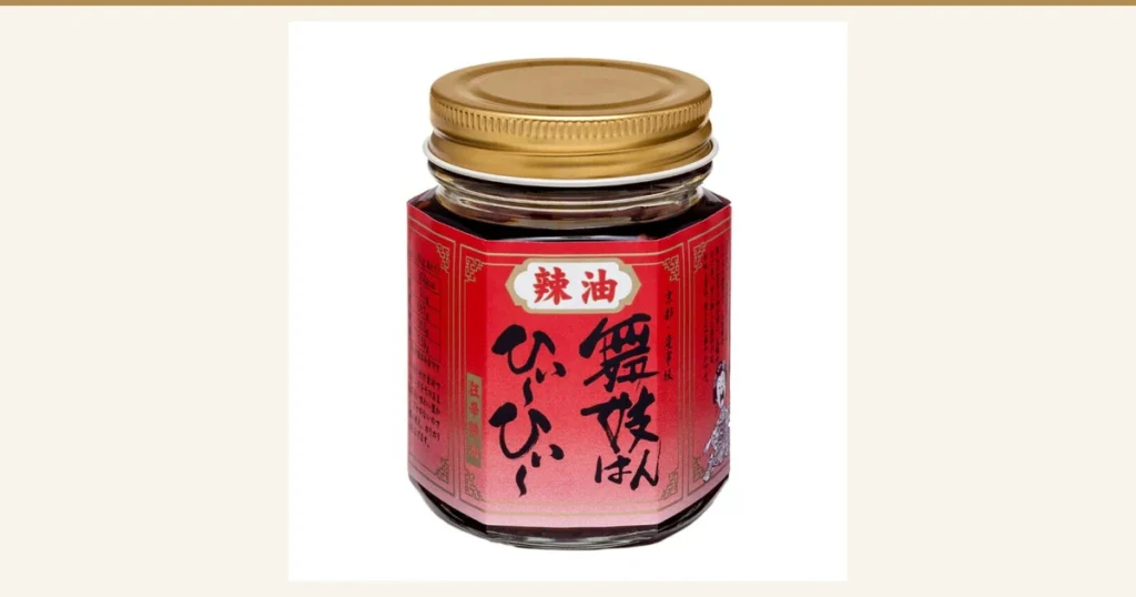 Product photo of one item from the 舞妓はんひぃ～ひぃ～ Set (Ichimi, Shichimi, Chili Oil): the chili oil, in a clear angular jar with a metal screw cap and a red label. Text printed on the label: “辣油”, “舞妓はんひぃ～ひぃ～”, “京都・産寧坂”. Small print and a small illustration at the edges of the label are too small to read