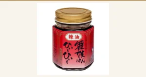 舞妓はんひぃ～ひぃ～セット（一味・七味・ラー油）のうち、ラー油の容器1点の商品写真。金属のねじ蓋をつけた角のある透明な瓶に赤いラベルが巻かれている。ラベルの印字「辣油」「舞妓はんひぃ～ひぃ～」「京都・産寧坂」。ラベルの両端には小さな印字とイラストがあるが、小さくて判読できない
