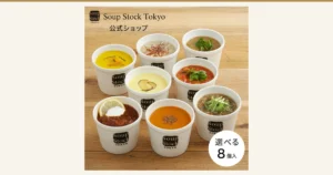 Product photo of the Soup Stock Tokyo choose-your-own 8-soup set. Eight soups in white cups, in colors such as yellow, pale yellow, orange, reddish brown, tomato red and brown, are lined up on a wooden table, topped with green onion, herbs, a lemon slice and red chili threads. The cups and toppings are a serving example; what the product contains is frozen soup packs. Text the store placed in the image: the “Soup Stock Tokyo” logo, “公式ショップ”, “選べる 8個入”