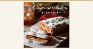 Product photo of a powdered-sugar-dusted stollen loaf with a few cut slices on a wooden board, with a red ribbon and a candle in the background (オーセントホテル小樽 Original Stollen in a Gift Box). A serving example in the cut state. Text added to the image by the shop: “Original Stollen”, “オリジナルシュトーレン”