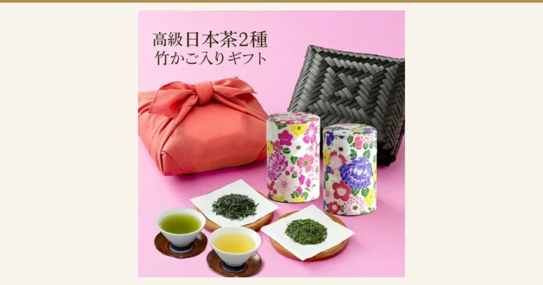 Product photo on a pink background showing a rose-colored furoshiki bundle, a black bamboo basket, two floral tea tins, tea leaves on paper and two cups of brewed tea (川本屋茶舗 autumn kuradashi new tea and Kakegawa tea bamboo basket set). Text the store placed in the image: “高級日本茶2種 竹かご入りギフト”. The brewed tea, teacups, saucers, and the paper and stands holding the tea leaves are not included in the product