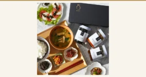 OKiNI 佃煮瓶3本セット 小倉屋山本のアイキャッチ。黒い専用化粧箱と、のり佃煮・雲丹ほたて・おかか明太の瓶3本を、ご飯・味噌汁・小皿の佃煮・トマトとチーズのサラダとともに上から撮った写真。ご飯・味噌汁・サラダ・器は盛り付けの例で、商品に含まれない。画像内の文字は瓶のラベル（「のり佃煮」「雲丹ほたて」「おかか明太」「OKINI」）のみ。