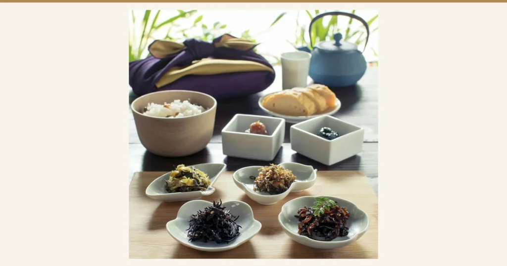 Serving-example photo of the Recommended Tsukudani Assortment (Kakuichi Yokocho): a purple and gold furoshiki-wrapped parcel behind six small plates and bowls of tsukudani, with rice, rolled omelet and a teapot also in the frame. The rice, omelet and teapot are not included in the product