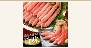 竹のざるに針葉樹の葉を敷いて殻つきの先端を残したずわいがにの棒肉を盛り、手前の黒い皿にも棒肉とライム、わさびを添えた商品写真(生本ずわいがに 棒肉ポーション)。画像内に店舗が画像に入れた表示として、「濃厚な甘みと旨味」の文字あり。ライム、わさび、針葉樹の葉は盛り付けの飾り。