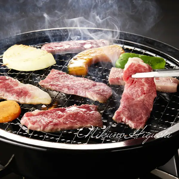Photo of a cooking example for Hida Gyu Karubi Yakiniku Mega Portion 1kg: beef for yakiniku, onion, pumpkin, green pepper, and carrot being grilled on a smoking grill grate, with a piece of meat held up with tongs. A cursive watermark of the shop name is at the bottom of the photo.