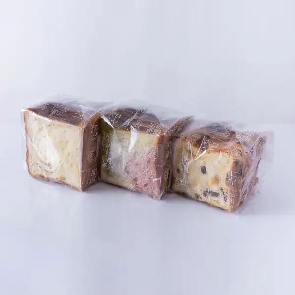 Product photo of 京都祇園ボロニヤ 元祖デニッシュ食パン (Ganso Danish Bread), three sliced 1-kin loaves: three loaves of Danish bread, each in its own clear bag, lined up side by side. From the left: plain, one with green, white and pink dough (apparently three-color strawberry), and raisin. The bags are printed in white with “京都祇園”, “ボロニヤ”, “GION BOLONIYA KYOTO” and a flower-pattern logo.