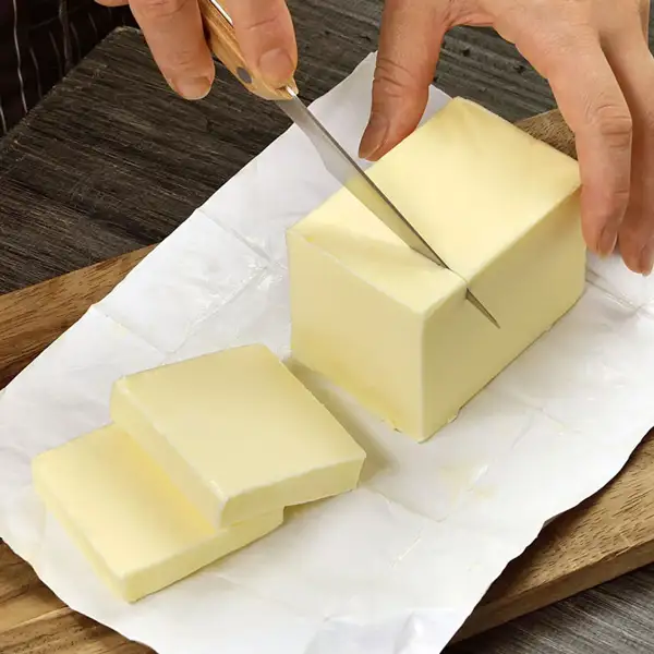 Hands cutting a block of Yotsuba Butter Unsalted on its wrapping paper with a knife, with two cut slices of butter in front.