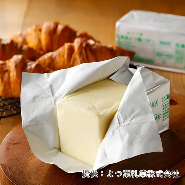 包み紙を開いて黄色いバターの塊を見せている、よつ葉バター 食塩不使用の商品写真。奥に同じ包装の箱とクロワッサン、手前に木の丸い板。右下に「提供:よつ葉乳業株式会社」の文字。