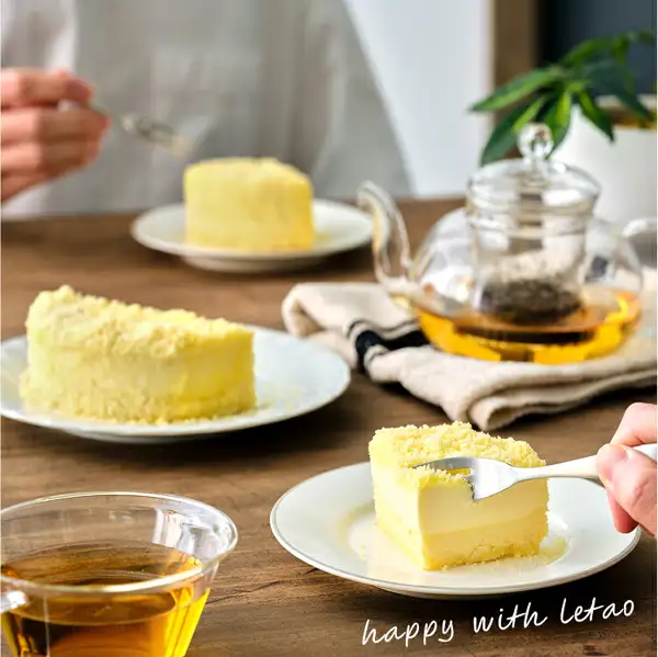 Serving example photo: on a wooden table are 3 white plates with cut slices of Fromage Double, a glass teapot, and a cup of black tea; a fork held in a right hand is scooping up the cheesecake. Text the shop placed in the image: "happy with letao"