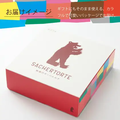 白い箱の上面に熊のイラストと「SACHERTORTE」「魅惑のザッハトルテ」の文字があり、側面が赤とカラフルな色の面で彩られた外箱の写真（果子乃季 魅惑のザッハトルテ 直径約15cm）。店舗が画像に入れた表示「お届けイメージ」「ギフトにもそのまま使える、カラフルで可愛いパッケージでお届け。」