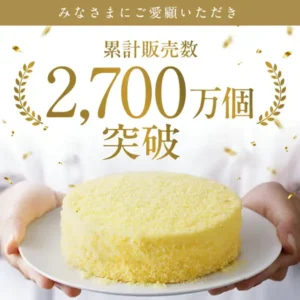 Product photo of LeTAO “Fromage Double” cheesecake topped with yellow crumbs, held on a white plate by two hands (no face visible). Text the shop placed in the image: “みなさまにご愛顧いただき” (“Thank you for your patronage”) and “累計販売数 2,700万個突破” (“Cumulative sales passed 27 million pieces”).