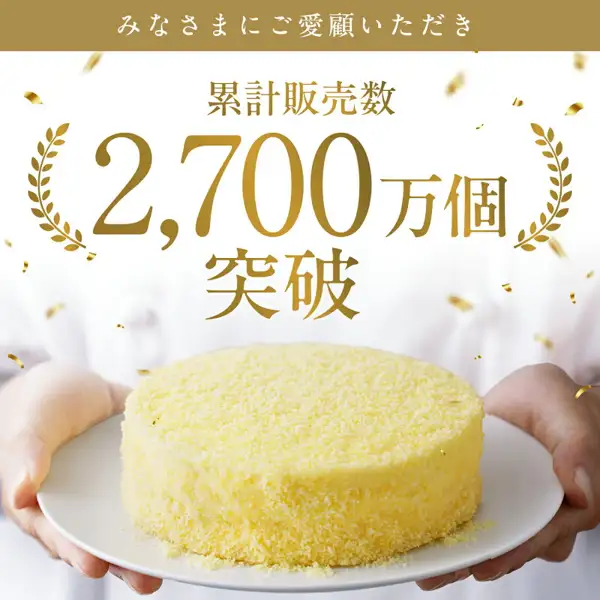 Product photo of LeTAO “Fromage Double” cheesecake topped with yellow crumbs, held on a white plate by two hands (no face visible). Text the shop placed in the image: “みなさまにご愛顧いただき” (“Thank you for your patronage”) and “累計販売数 2,700万個突破” (“Cumulative sales passed 27 million pieces”).