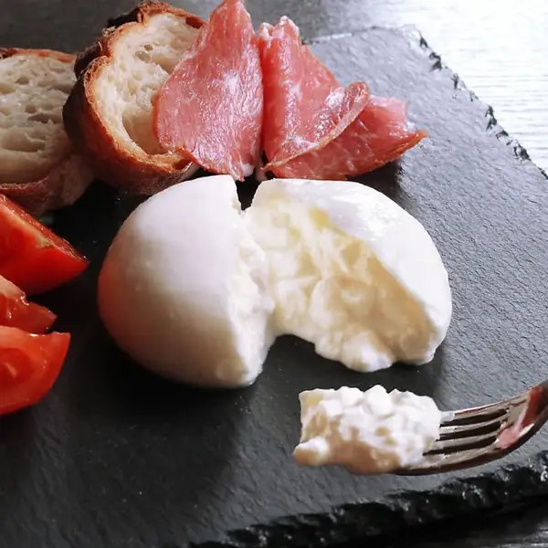 黒いスレート皿の上で割った白い丸形のブラータ（生モッツァレラ）チーズと、フォークですくった中身が写っている商品写真（花畑牧場 ブラータ〜生モッツァレラ〜 チーズ 70g×12個）。皿には生ハム・バゲットの薄切り・くし切りのトマトも並んでいるが、これらは商品に含まれない盛り付け例