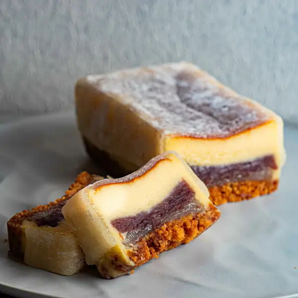 Product photo of a baked cheesecake wrapped in habutae mochi on a white plate: a block at the back and cut pieces in front, whose cross-section shows layers of cheese batter, sweet red bean paste and a crumble base (photo of the cake after cutting). RUNNY CHEESE 羽二重ベイクドチーズケーキ 16cm (habutae baked cheesecake)