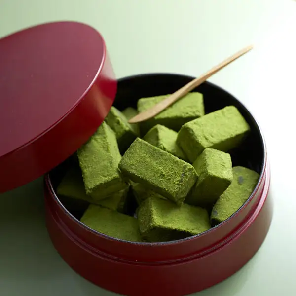 Photo of square nama chocolates coated in matcha powder, served in a round red container with its lid off and a bamboo pick (serving example). Itohkyuemon Uji Matcha Nama Chocolate, 16 Pieces, Matcha Flavor