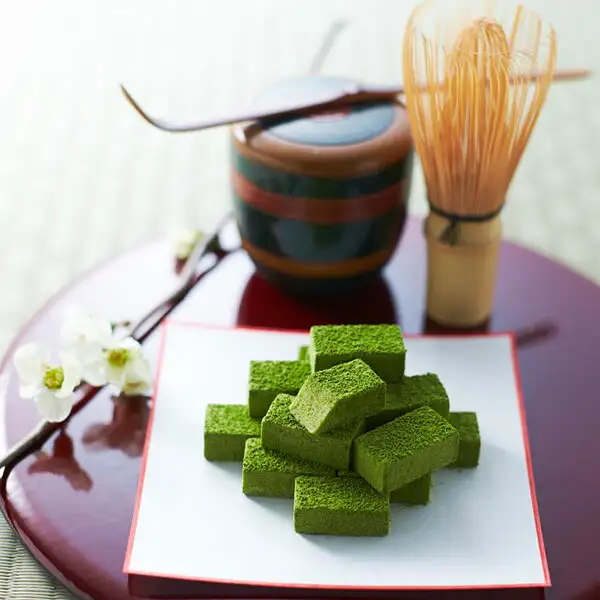 赤い丸盆にのせた白い懐紙の上に、抹茶の粉をまとった角形の生チョコを積んだ写真（盛り付け例）。奥に棗・茶杓・茶筅、左に白い花の枝。宇治抹茶 生チョコ 16粒 抹茶味（伊藤久右衛門）