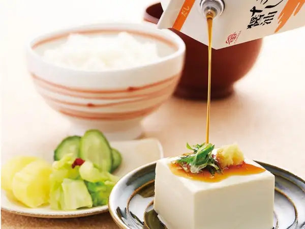 鎌田醤油 だし醤油の盛り付け例の写真。オレンジの帯に「だし」の文字と「KAMADA」のロゴが入った紙パックから、器に盛った豆腐（刻んだ青い薬味とおろし生姜をのせた冷ややっこ）にだし醤油を注いでいる。奥に白いご飯を盛った茶碗と茶色の汁椀、左手前に黄色い漬物・きゅうり・キャベツを盛った小皿。紙パックの容量は画像からは判別できない