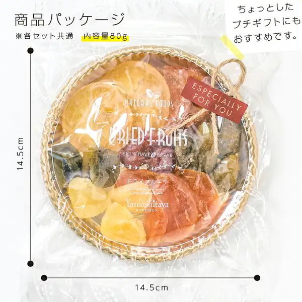 透明な袋に入った丸い編みかご風のトレイに、輪切りのドライフルーツや黄色い角切りなどが円形に盛られた、国産ドライフルーツ 食べ比べ7種（定番セット）山下屋荘介の商品パッケージ写真。トレイに麻ひもと赤い札が付き、袋の左右と下に寸法線がある。店舗が画像に入れた表示「商品パッケージ」「※各セット共通 内容量80g」「ちょっとしたプチギフトにもおすすめです。」、寸法「14.5cm」「14.5cm」。袋の印字「NATURAL FOODS」「DRIED FRUITS」「ドライフルーツ」「Yamashitaya KITCHEN」、札「ESPECIALLY FOR YOU」。