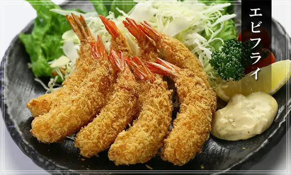 Serving-example photo of ebi fry (breaded fried shrimp) on a black plate, served with shredded cabbage, lettuce, parsley, cherry tomatoes, lemon and tartar sauce (Kouragumi Wild-Caught Ebi Fry, 8 Pieces). Text the shop placed in the image: “エビフライ” (ebi fry)