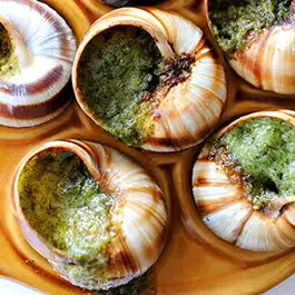 Photo of baked shell-on escargot arranged in the hollows of a plate. Green butter fills the spiral, striped shells. Introduction image (cooking example) for ハイ食材室's “Escargot à la Bourguignonne from France, Shell-On with Butter, 30 Pieces”. No text is baked into the image.