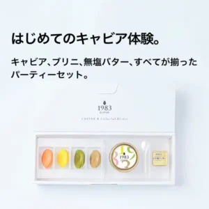 宮崎キャビア 1983 (12g) & ブリニ セット的商品照片：白色礼盒中摆放着橙、黄、绿、茶4种颜色的4片ブリニ、盖子为白色带金边、印有「1983」的鱼子酱瓶、贝壳勺和金色包装的黄油。图片内为店铺自行加入的文字：「はじめてのキャビア体験。」「キャビア、ブリニ、無塩バター、すべてが揃ったパーティーセット。」。盒子和瓶盖上印有「1983 J.CAVIAR」「CAVIAR & Colorful Blinis」。