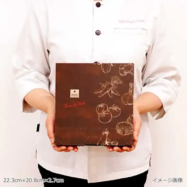アスザックフーズ 20品目の野菜ときのこが摂れるスープ食べ比べセット（6食）の専用の箱を、白いコックコートを着た人が胸の前で両手で持った写真（顔は写っていない）。焦げ茶色の箱に「ASUZAC FOODS」のロゴと赤い筆記体の「Farm to Fork」、人参やトマトなどの野菜の線画がある。コックコートの胸に「SHOP Chef’s Table」の文字。画像内に店舗が画像に入れた表示として、「22.3cm×20.8cm×2.7cm」「イメージ画像」の文字あり。
