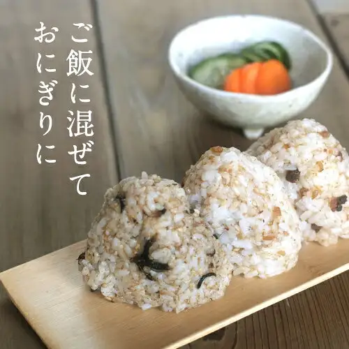 Serving-example photo for the Furikake Set of 4, Additive-Free (Suzuki Katsuobushi-ten 鈴木鰹節店): three rice balls speckled with what looks like hijiki and sesame sit on a long, narrow board on a wooden table, with a white bowl of sliced cucumber and carrot in the back. Text the shop placed in the image reads “ご飯に混ぜて おにぎりに” (Mix it into rice for rice balls).