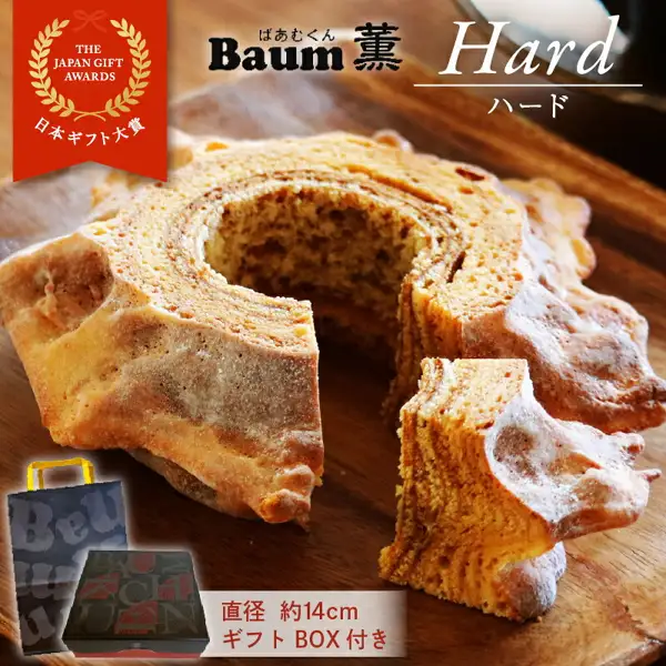Product page image for the furusato nozei gift “ばあむくん かため” (Baamukun Katame). On a wooden board, a cross-section of a tree-stump-shaped baumkuchen shows ring-like layers; one cut slice is at the lower right, and a black gift box and a black paper handbag are at the lower left. Text the shop placed in the image: “THE JAPAN GIFT AWARDS”, “日本ギフト大賞” (Japan Gift Awards), “Baum薫”, “Hard ハード” (hard), “直径 約14cm” (diameter about 14cm), “ギフト BOX付き” (comes with a gift box).